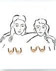 On a white background is a gold pair of boob shaped stud earrings on a white card packaging with an illustration of two women.
