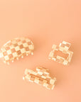 On a peachy background is three different shaped claw clips with a white and clear checker print. One clips is a slightly smaller square shape, the other is a rectangular shape and the third has a rounded edge.