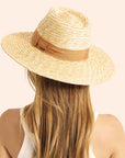 On a cream background is a model wearing a light honey colored straw sun hat with a tan ribbon around the base.