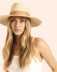 On a cream background is a model wearing a light honey colored straw sun hat with a tan ribbon around the base.