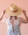 On a cream background is a light honey colored straw sun hat with a tan ribbon around the base.