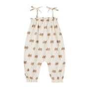 On a white background is a children's ivory jumpsuit with a neutral tan palm tree pattern and tie shoulder strap details. 