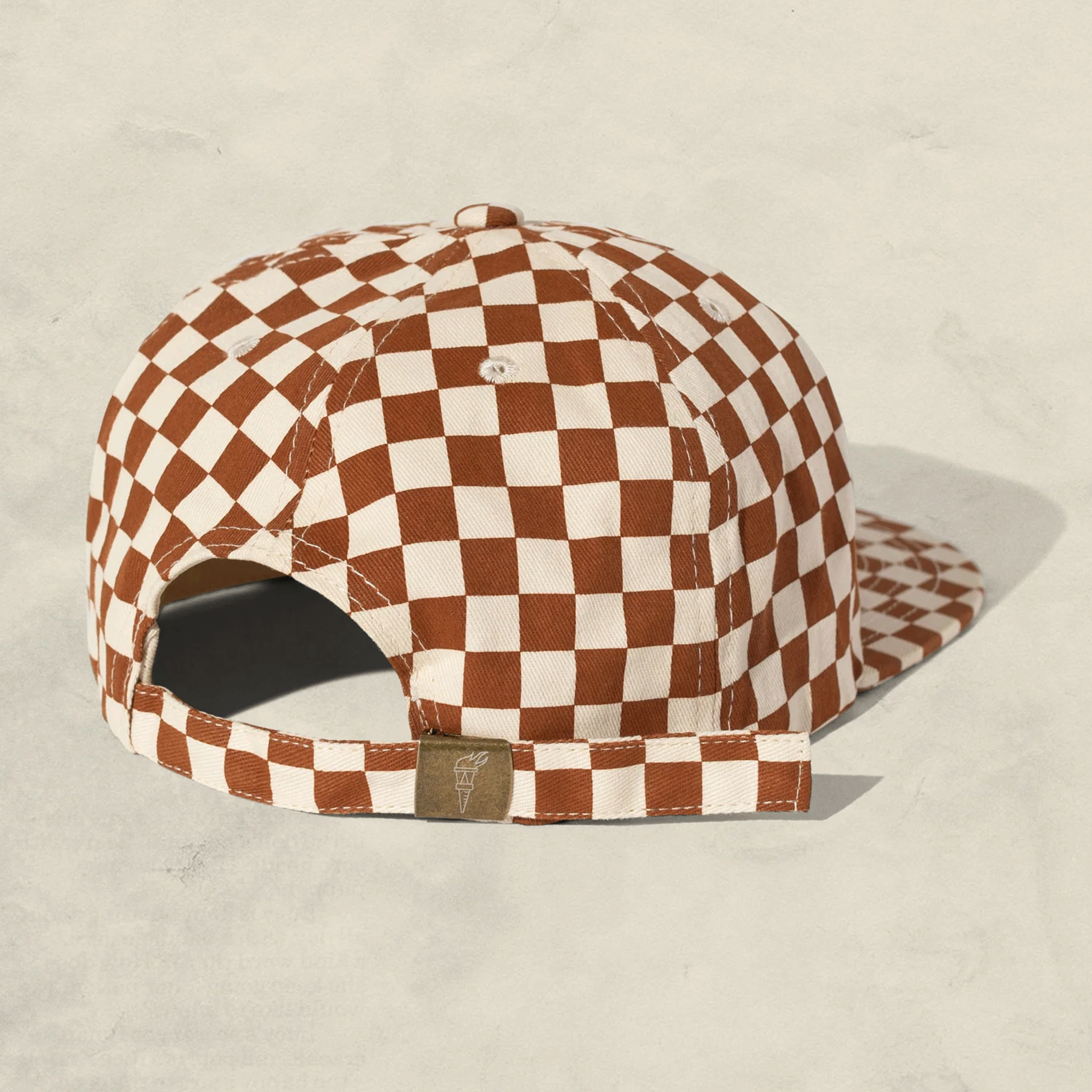 On a grey background is a rust and ivory checker pattern baseball hat for kids with a mental adjustable back. 