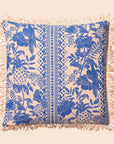On a cream background is a blue and ivory tropical print pillow with a tassel detail along the edges.