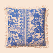 On a cream background is a blue and ivory tropical print pillow with a tassel detail along the edges.