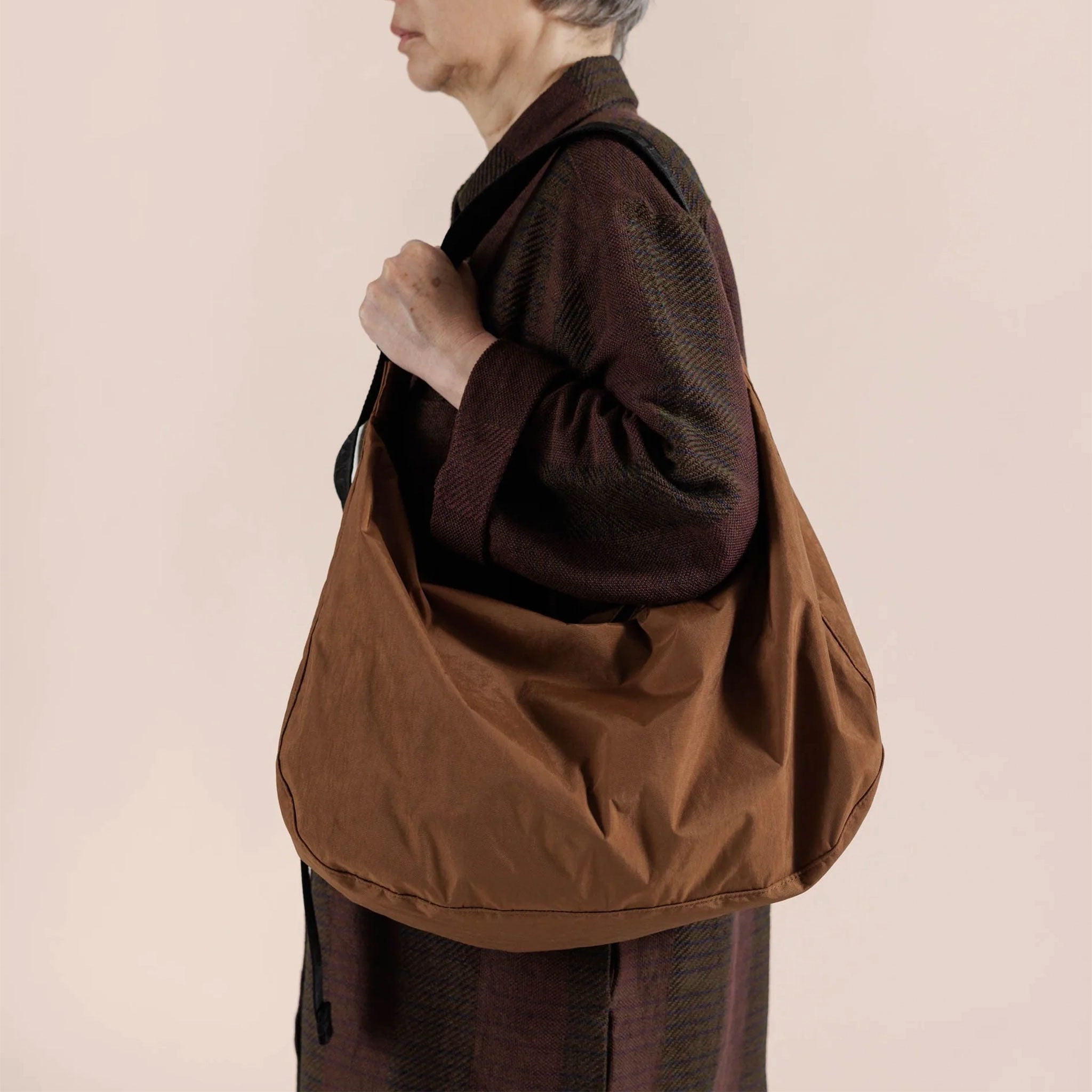 On a peachy background is a model wearing a brown crescent shaped nylon bag with a black strap. 