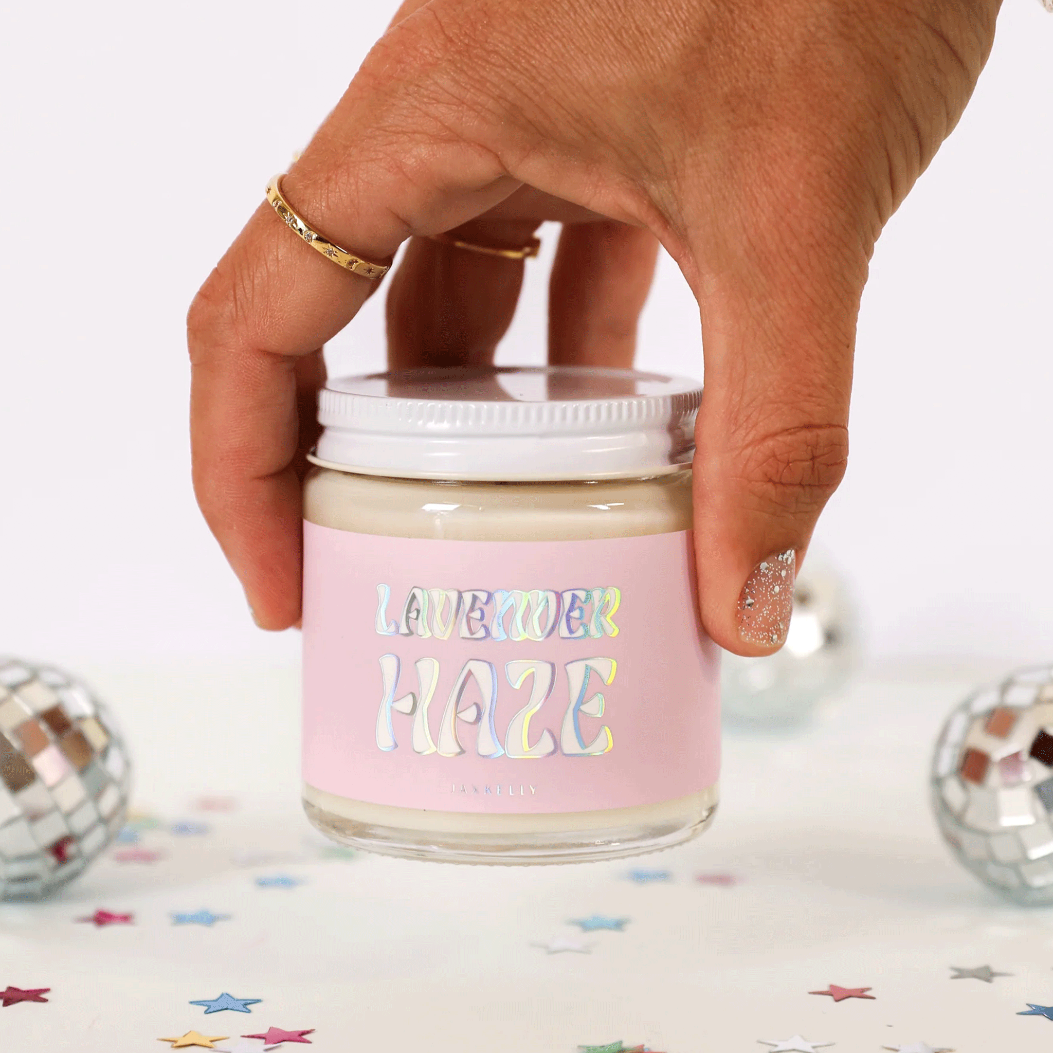 On a white background is a model's hand holding the 4oz glass jar candle with a light pink label that reads, "Lavender Haze" in a holographic wavy font. 