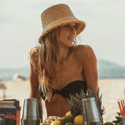 A woven bucket hat in a light straw color with a frayed edge detail along the edge of the brim and a tiny hemlock hat metal emblem in the center.