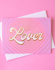 On a pink background is a pink and purple gradient card with gold foiled heart shaped outlines and ivory text that reads, "Lover".