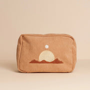 On a tan background is a peachy, tan corduroy makeup bag with a minimalist design of a desert mountain range along with the sun and moon above it.