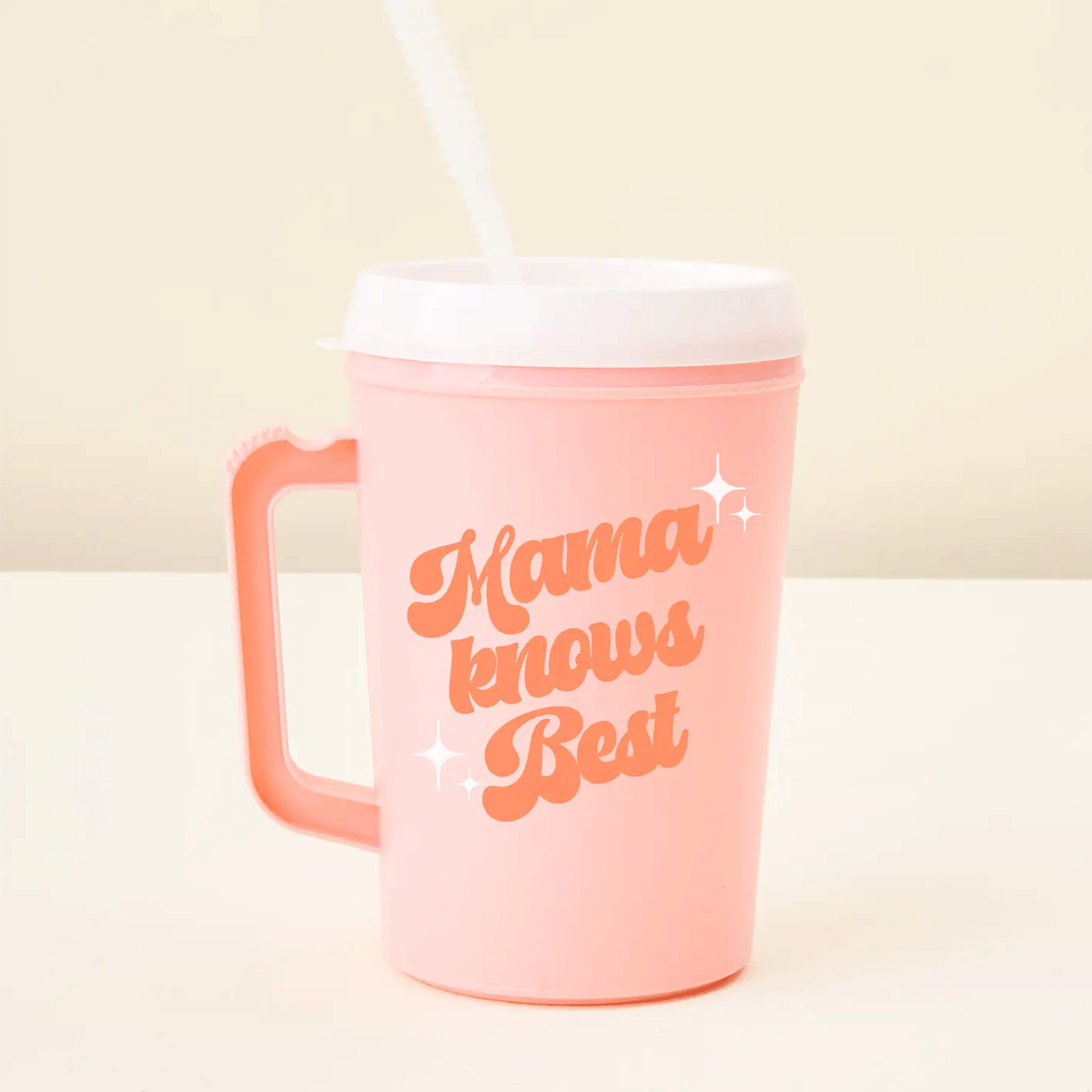 On a tan background is a pink travel mug with a handle and a white lid and straw and text on the front that reads, &quot;Mama knows Best&quot;. 