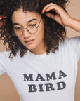 Woman in an off-white "MAMA BIRD" t-shirt and glasses, touching dark curly hair, with a beige background.