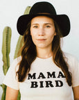 Woman in an off-white "MAMA BIRD" t-shirt and black fedora hat, standing in front of a cactus.