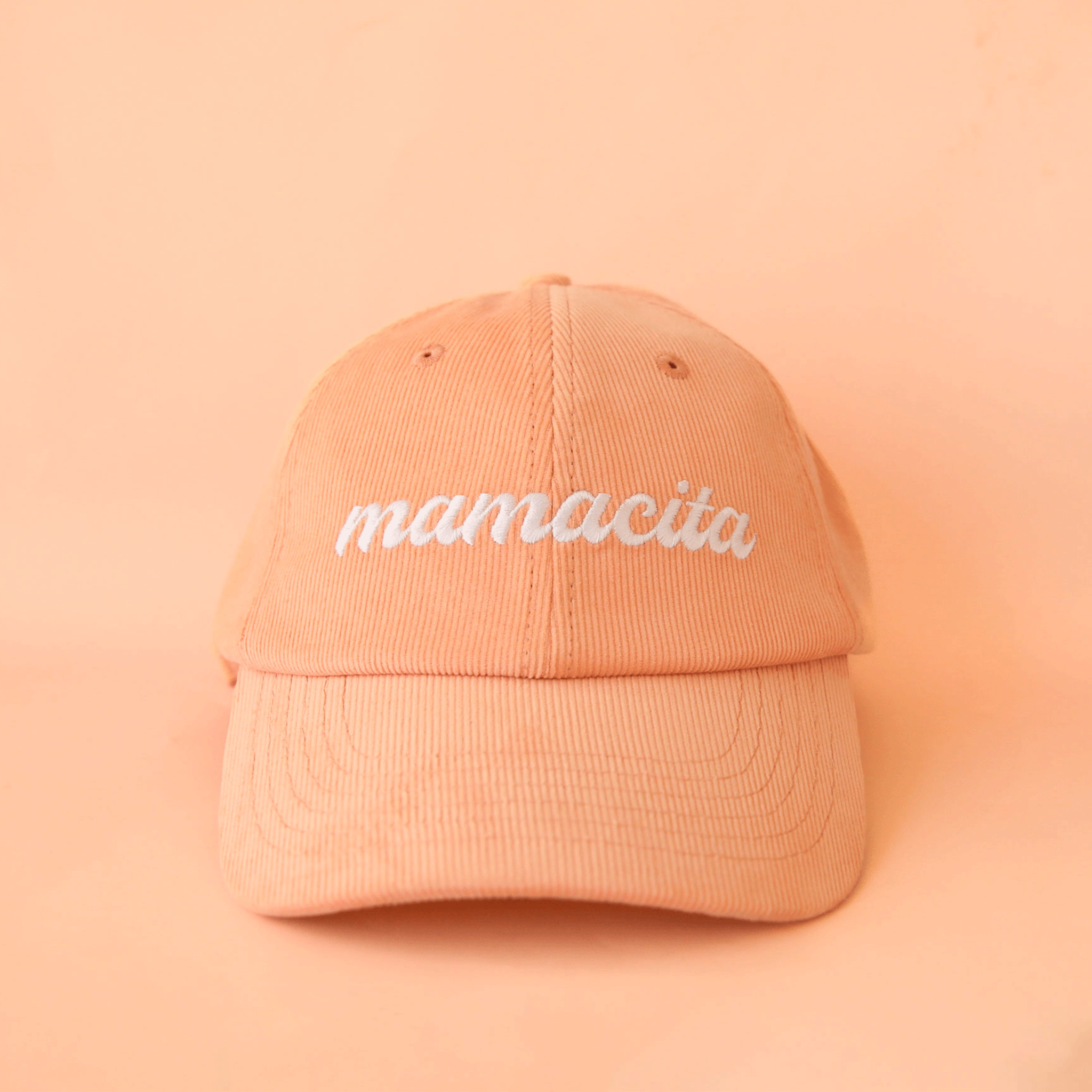 On a peach background is a peach colored corduroy baseball hat with white embroidery that reads, &quot;mamacita&quot;.
