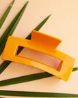 On a peachy background is the Havana Claw Clip in the color Mango which is a yellowy orange shade with a rectangular shape.