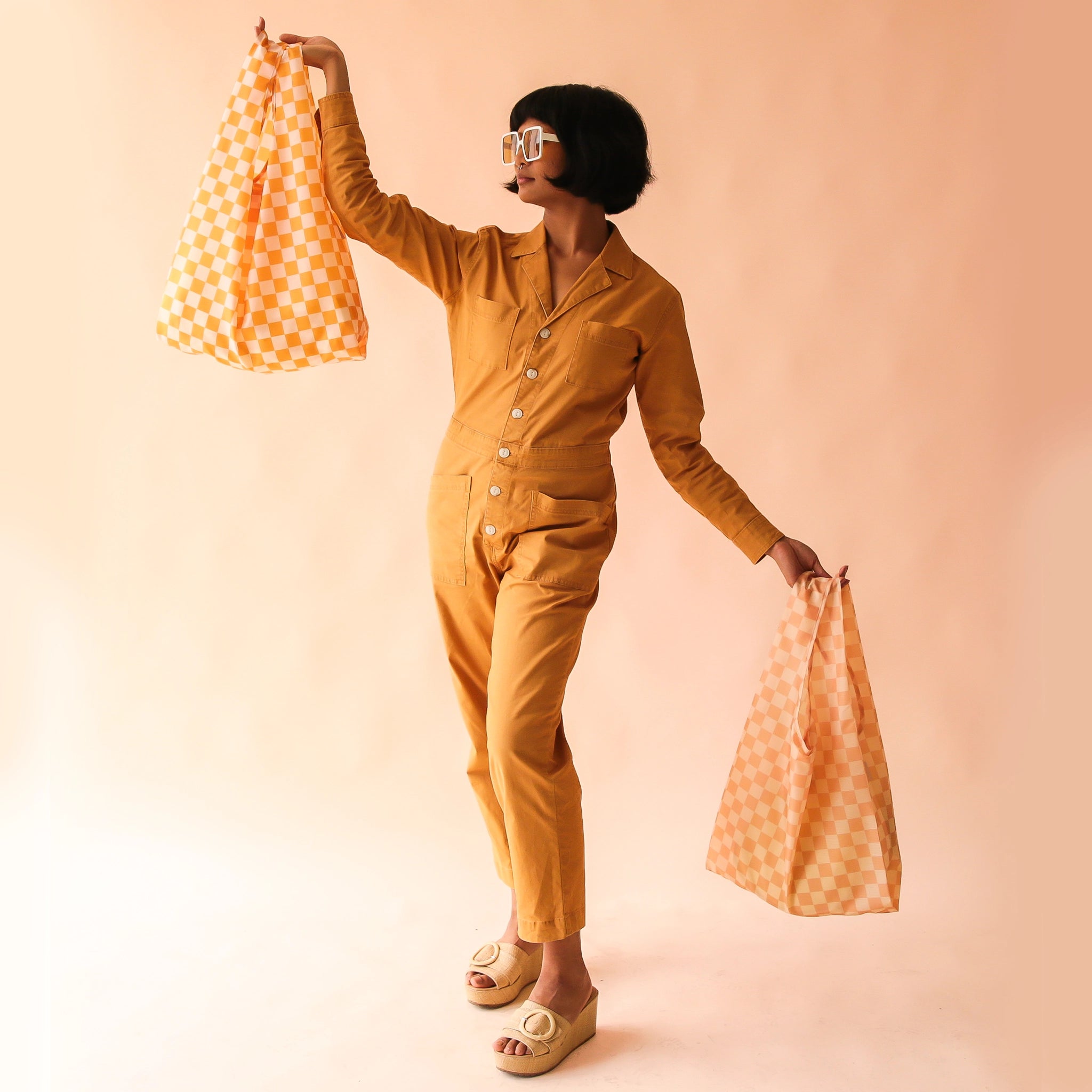 On a tan background is a mode holding two of our reusable bags one in marigold. 