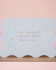 On a pink background is a wavy edged light blue card with gold foiled text that reads, "have yourself a merry little Christmas".