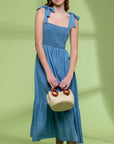 On a green shadow background is a model wearing a blue flowy midi dress with tie shoulder straps and a square neckline.