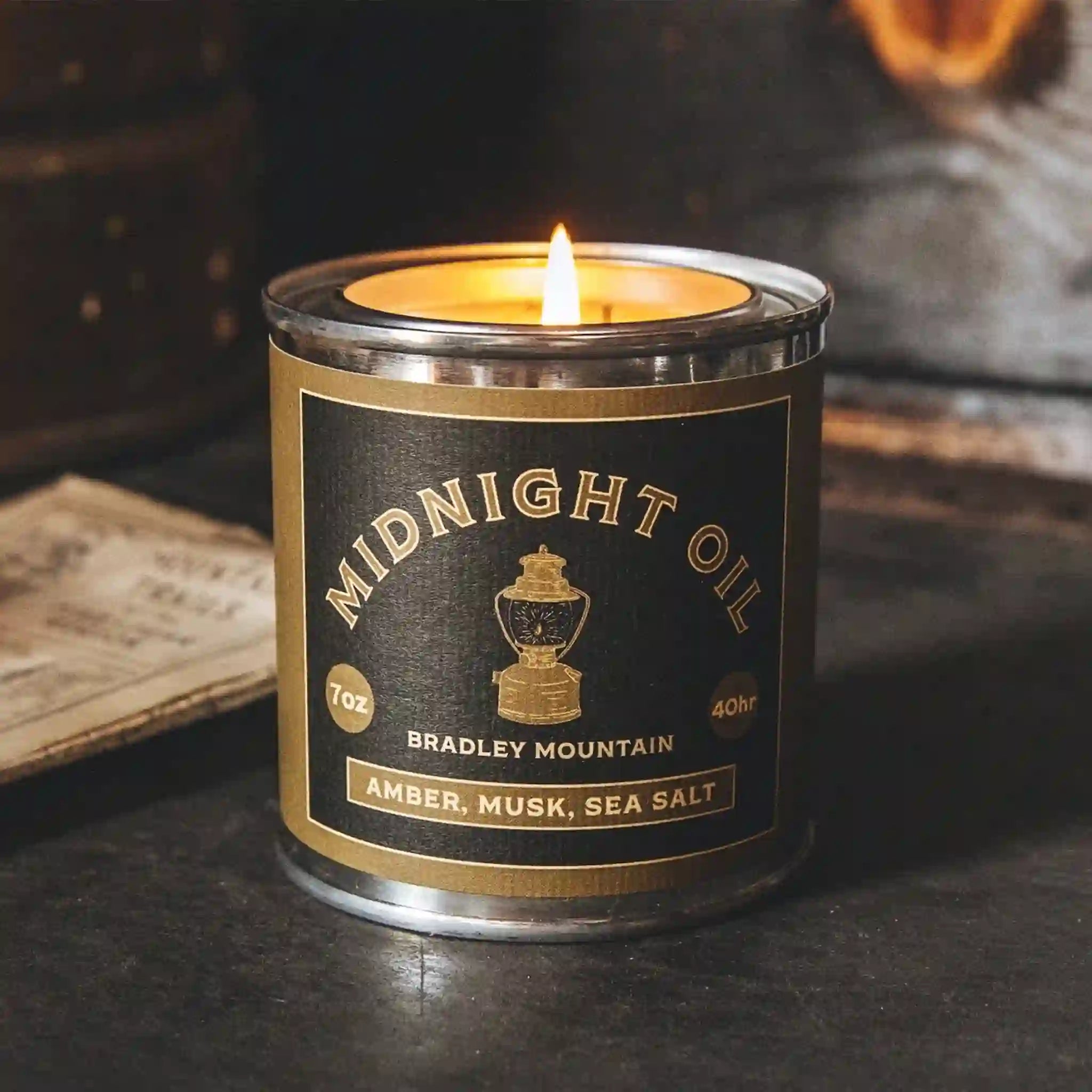 On a grey background is a green and black tin candle with arched text that reads, &quot;Midnight Oil Bradley Mountain&quot;.