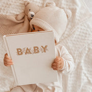 A model baby holding a neutral ivory colored book with gold lettering on the front that reads, "Baby".