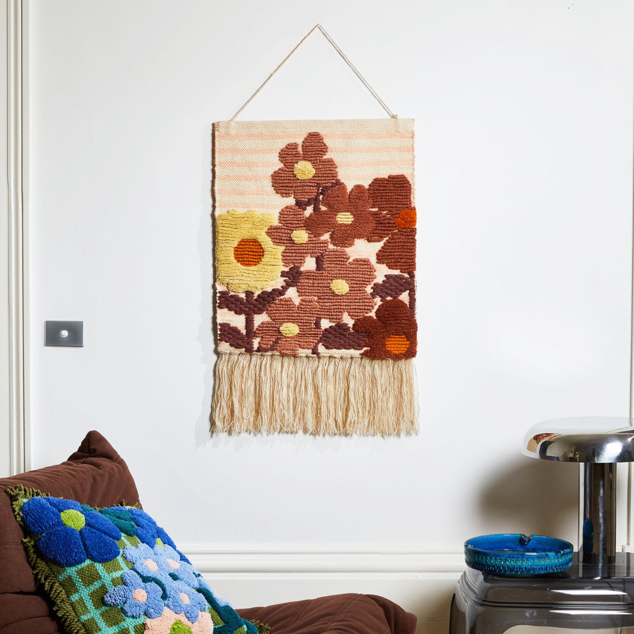 A hanging piece of woven wall art featuring rust and yellow colored floral prints and a fringe detail hanging from the bottom edge. 