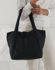 On a white background is a model holding the Mini Cloud Bag in the shade black. It is a rectangular shaped puffy bag with shoulder straps and a wide opening.