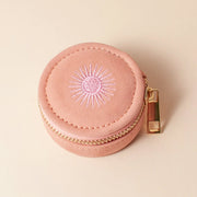 A rose colored circular jewelry case with a gold zipper and a sun design.