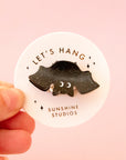 A black speckled bat shaped mini hair clip on a round piece of packaging that reads, 'Let's Hang Sunshine Studios'.