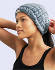 A fuzzy light blue extra wide headband folded up in its packaging that has a white label and black text that reads, "Extra Wide Spa Headband".