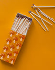 A rectangle box of matches with a repeating red mushroom design along all edges besides the top and bottom. The matches that are inside are also photographed here. They are wooden matches the same length as the box with a red striking tip for lighting.