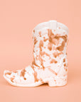 On a peachy background is a brown and white ceramic cowboy boot shaped candle.