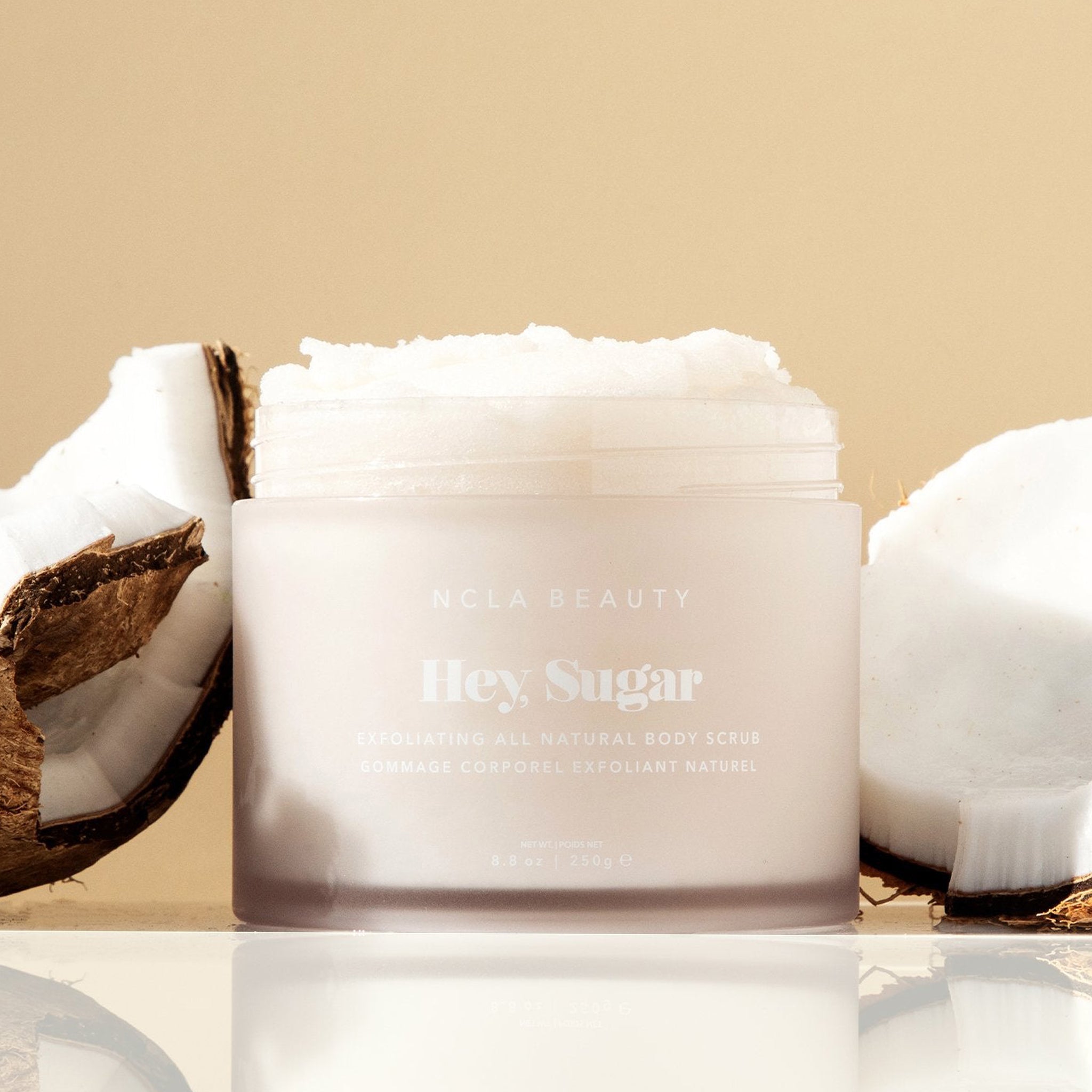 On a tan background is a container of white colored body scrub with text on the front that reads, &quot;Hey, Sugar&quot;.