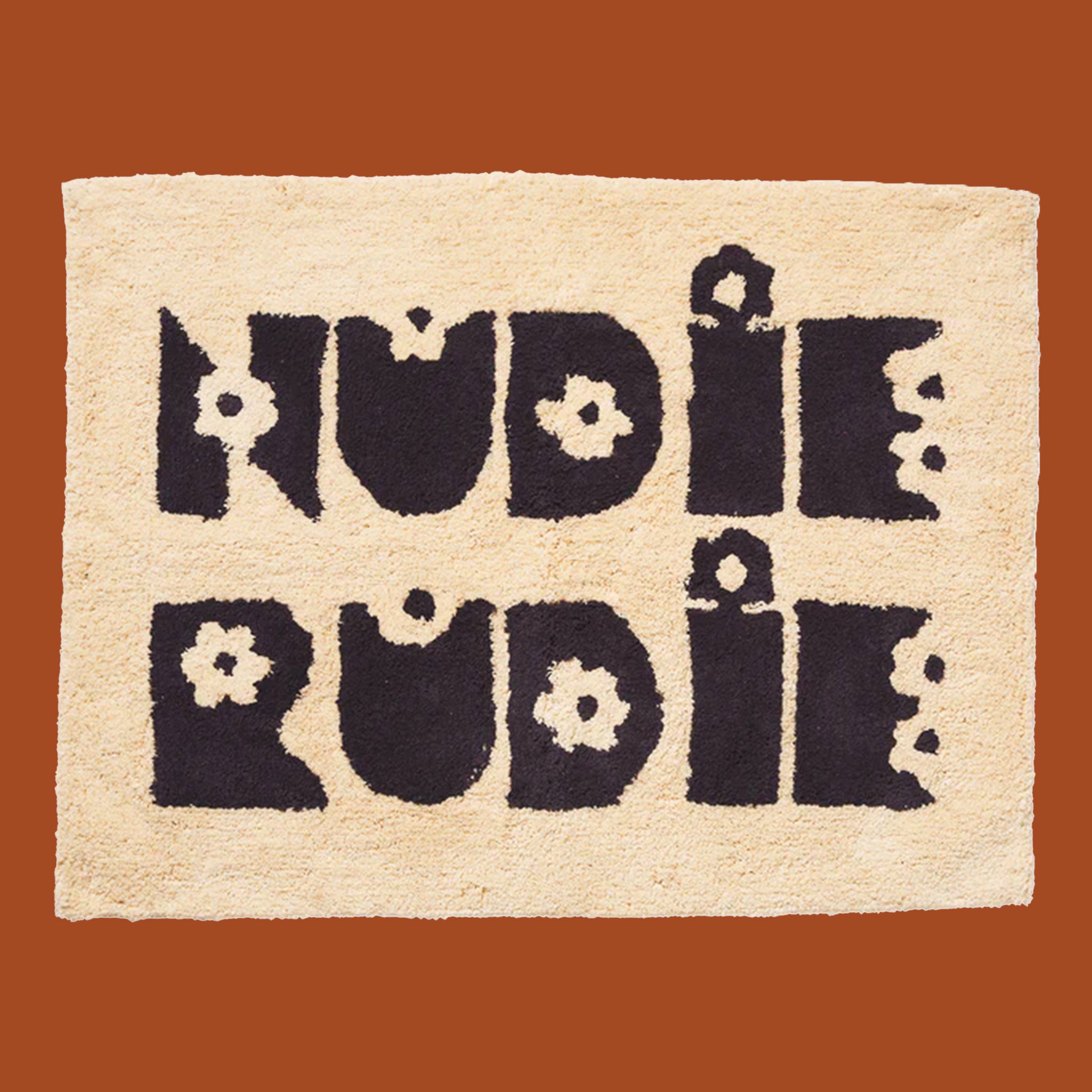 On a burnt orange background is a tan bath mat with black text that reads, "Nudie Rudie".