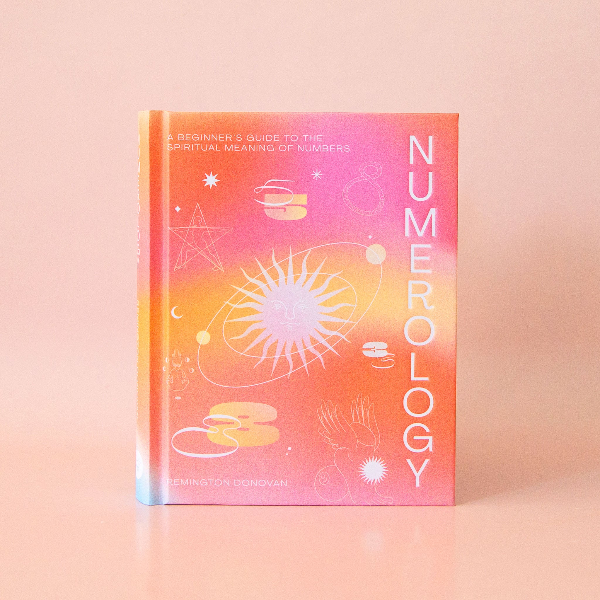 On a peachy background is a hot pink and orange book with a white title running vertically down the right side that reads, "Numerology", "A Beginner's Guide To The Spiritual Meaning of Numbers".