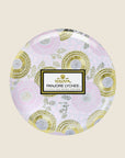 Inside the tin is a white candle with three white wicks. Leaning against the right side is a round tin lid. The lid has a light pink and gold floral pattern on it. In the middle is a white sticker with gold and black text inside.