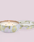Inside the tin is a white candle with three white wicks. Leaning against the right side is a round tin lid. The lid has a light pink and gold floral pattern on it. In the middle is a white sticker with gold and black text inside.