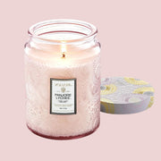 A light pink glass jar with a short tapered top. The jar has an etched in floral pattern around it. In the middle is a white sticker with a black and gold border. At the top is gold text that reads ‘Voluspa’ and black text that reads ‘Panjore Lychee.’ Leaning against the right side of the jar is a round tin lid. The lid is light pink with a gold floral pattern.