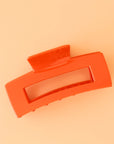 On a peachy background is the Havana Claw Clip in the color Papaya which is a bright reddish orange shade with a rectangular shape.