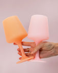 A pair of silicone wine glass shaped cups in a peach shade and a light pink shade. Photography props not included with purchase.