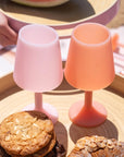 A pair of silicone wine glass shaped cups in a peach shade and a light pink shade. Photography props not included with purchase.