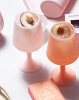 A pair of silicone wine glass shaped cups in a peach shade and a light pink shade. Photography props not included with purchase.