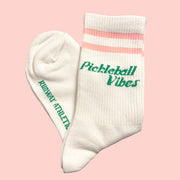 On a light pink background is a pair of white socks with pink stripes and green text that reads, "Pickeball Vibes".