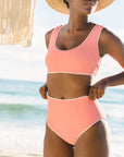 A model wearing a 1960's inspired swim suit with a wavy edge detail and high waist bottoms. The color of the swim suit is a coral pink hue.