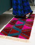 A pink, red, rust and teal triangle patterned bath mat with tassel details on the two ends.