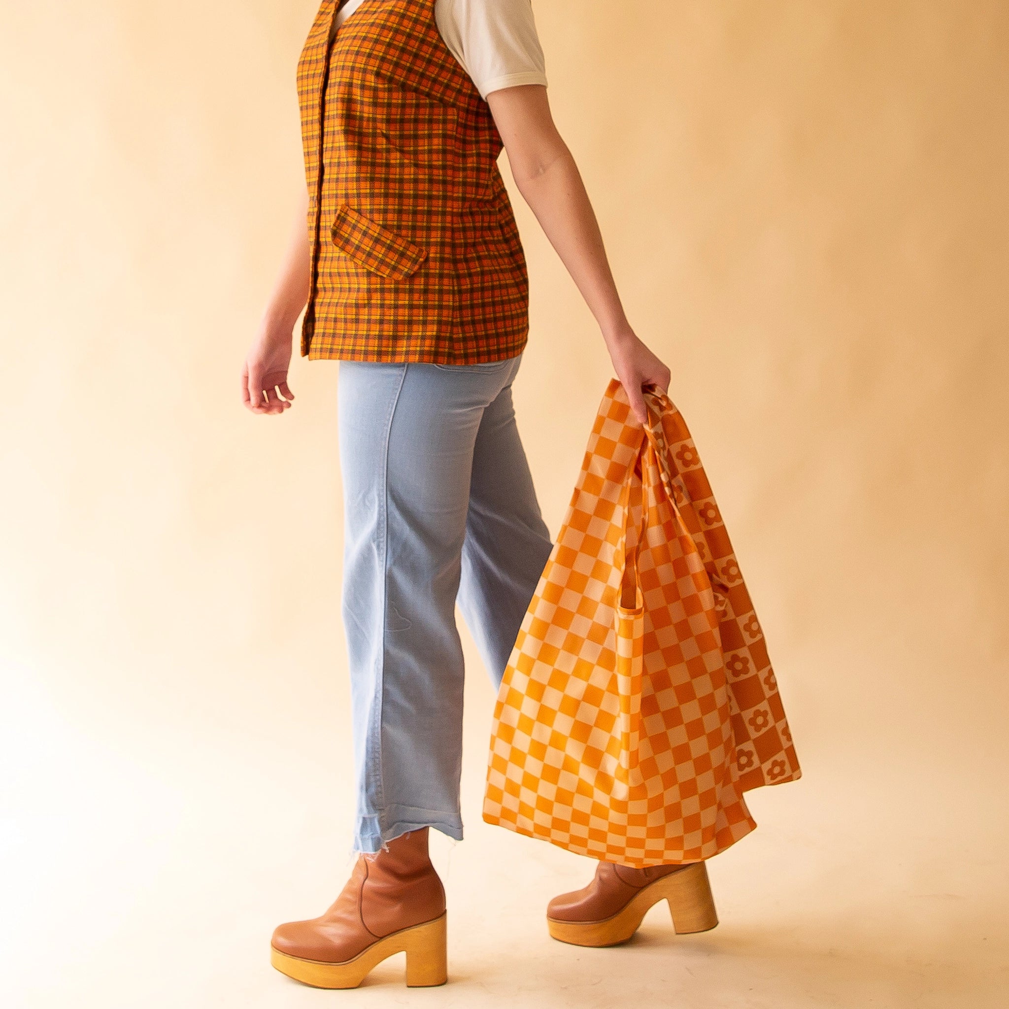 On a light peach background is a model holding two reusable nylon bags. One is a dark and light orange checkered print and the other is a burnt orange and light orange checker print with flowers in the squares. 