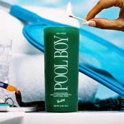 A thin clear glass candle with a green wax candle inside and has white text that reads, "Pool Boy".