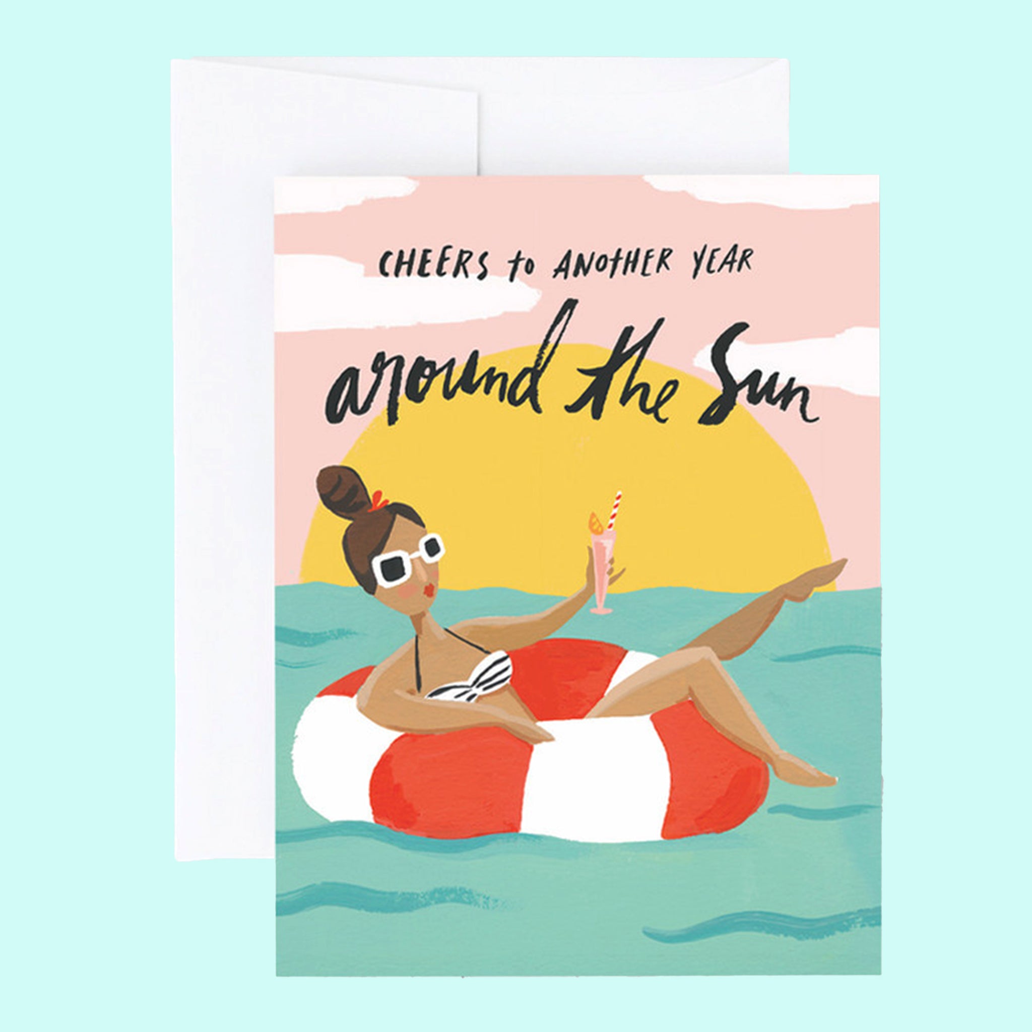 On top of a white envelope is a card. The card is a drawing of a blue ocean with a pink sky and yellow setting sun. In the pink sky is black text that reads ‘cheers to another year around the sun.’ In the water is a red and white striped inner tube with a girl laying inside. She is wearing white sunglasses and holding a pink drink.