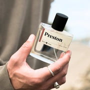 A clear glass bottle of cologne with a black wooden cap with a label that reads, 'Preston'.