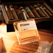 A clear glass bottle of cologne with a black wood cap and a label that reads, 'Primo'.