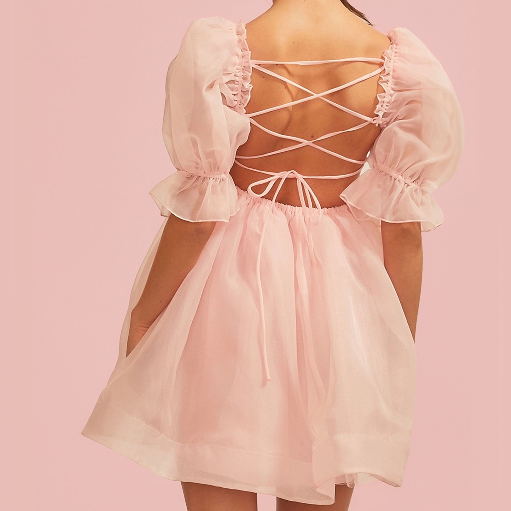 On a pink background is a model wearing a light pink puff sleeve dress with a tie up back. 